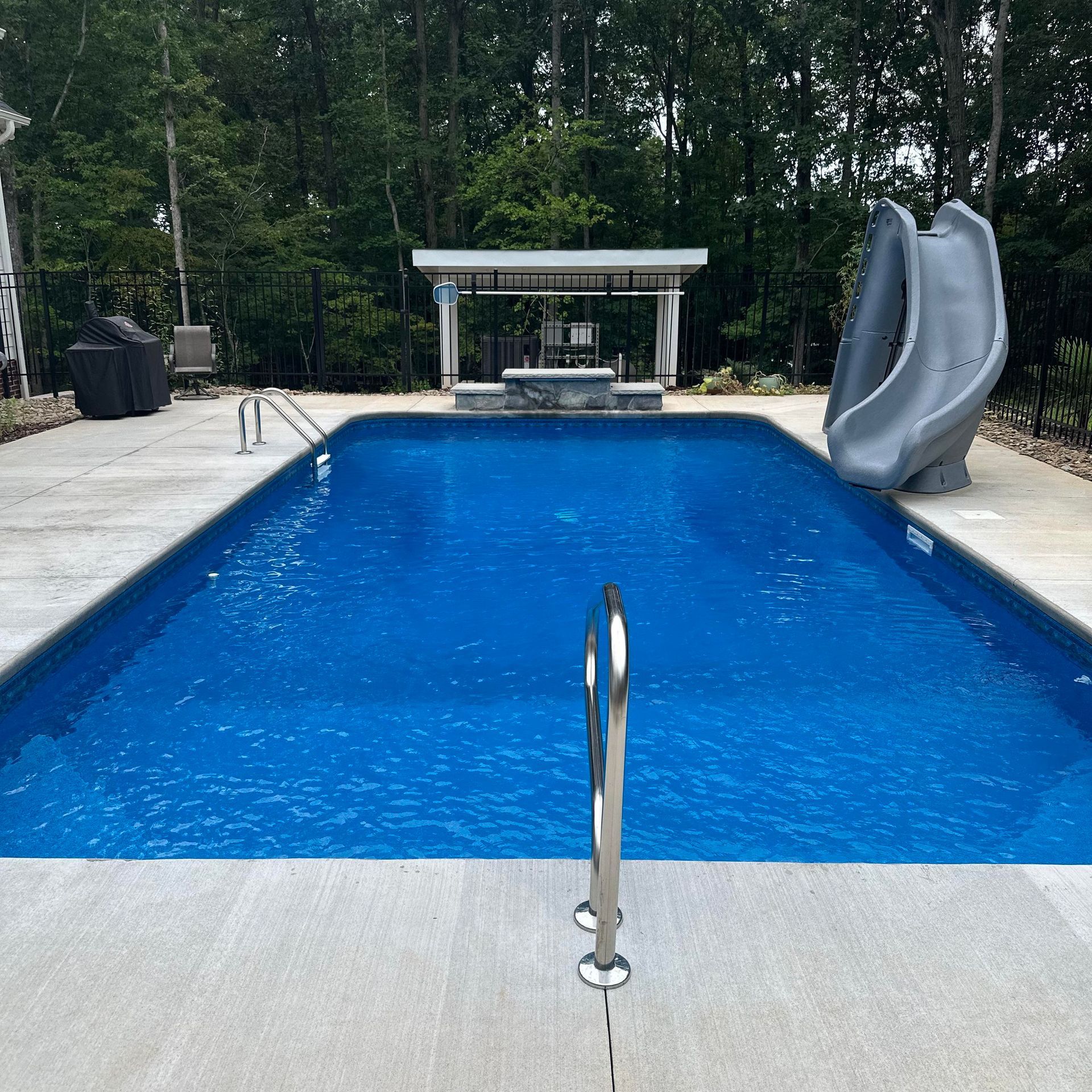 Backyard swimming pool with blue water and a gray slide, surrounded by a concrete deck and trees.