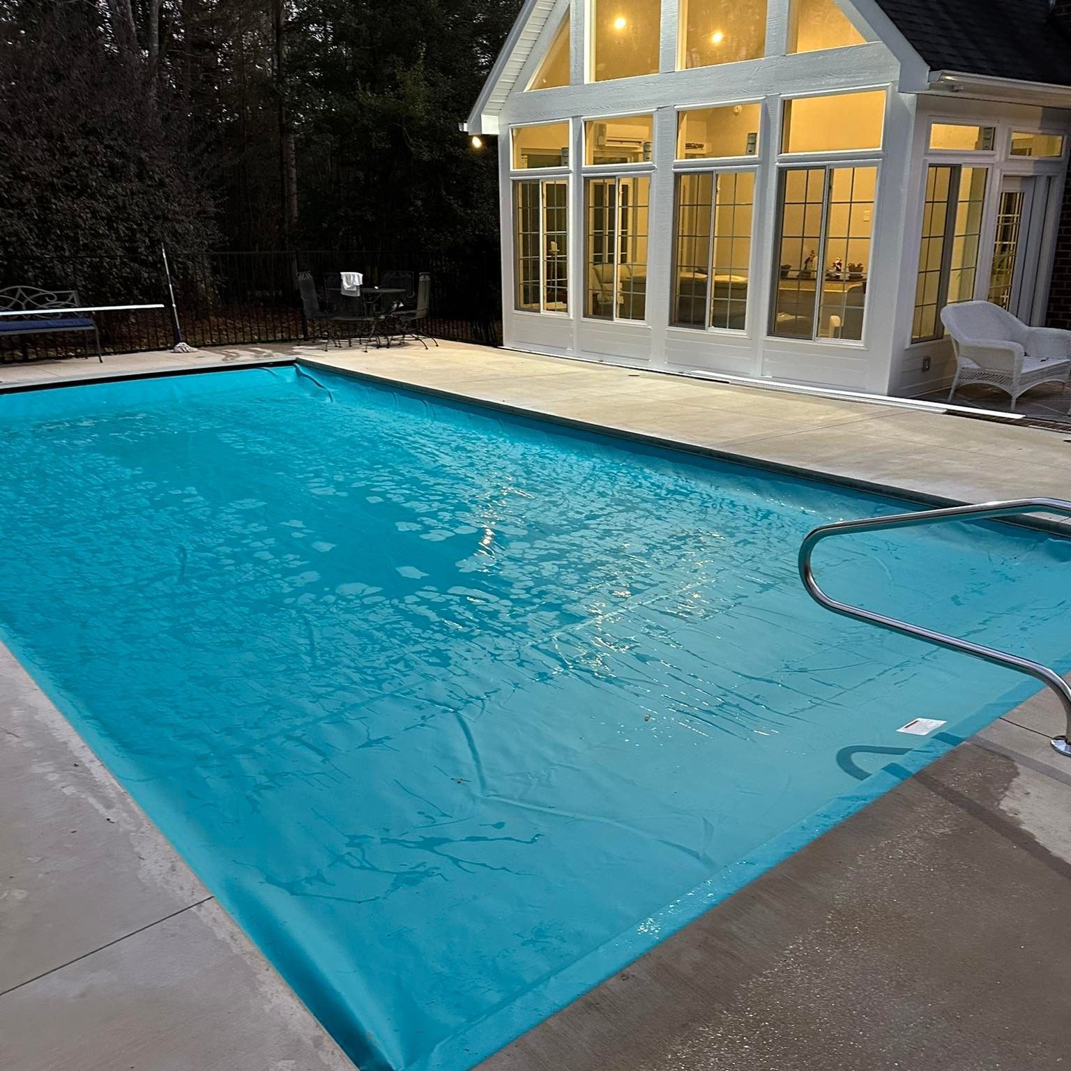 Backyard swimming pool beside a lit glass sunroom at dusk, with a pool hose in the water.