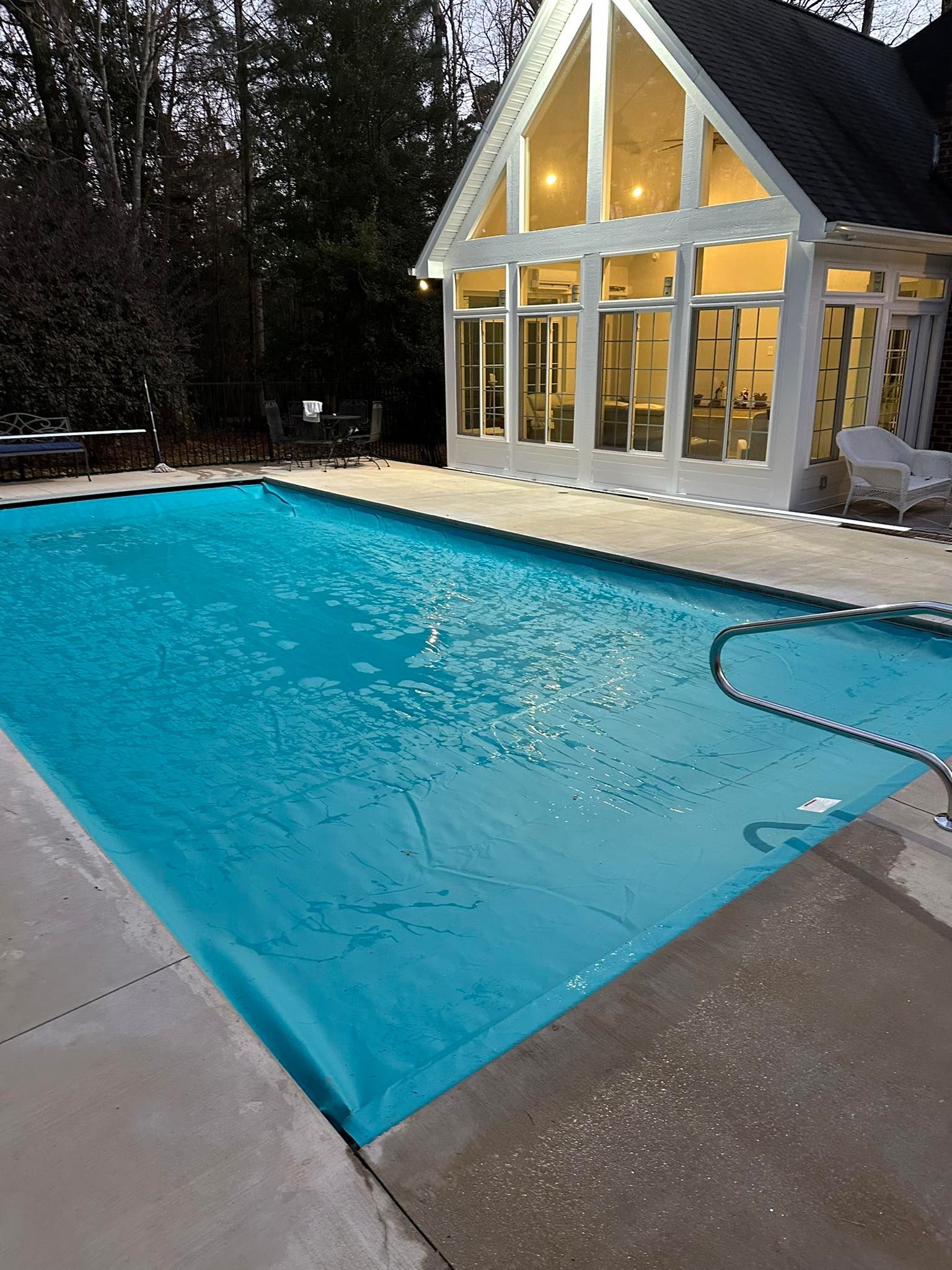 Rectangular backyard pool with turquoise cover beside a lit modern house at dusk