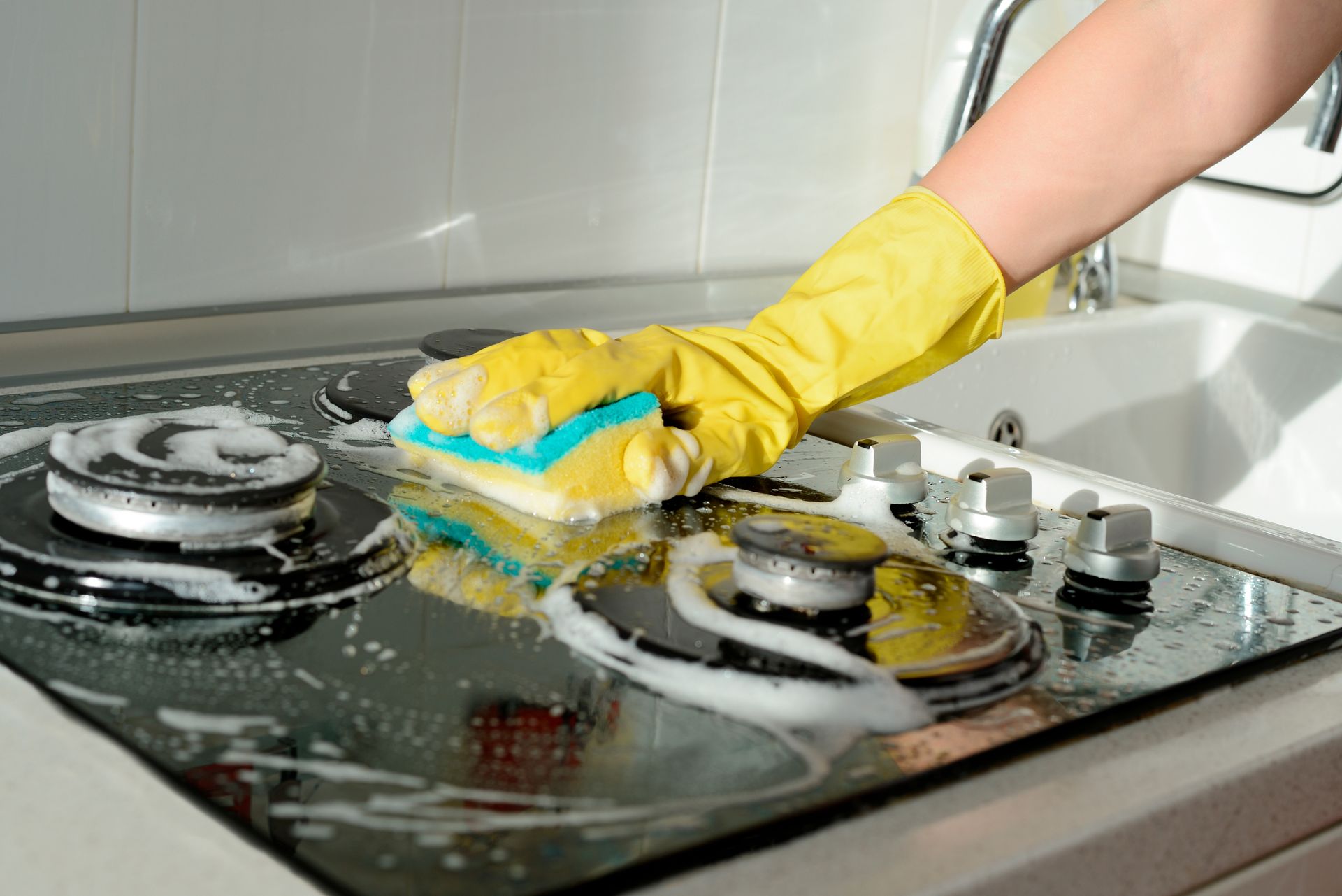 Hand in yellow glove scrubbing a dirty glass stovetop with sponge.