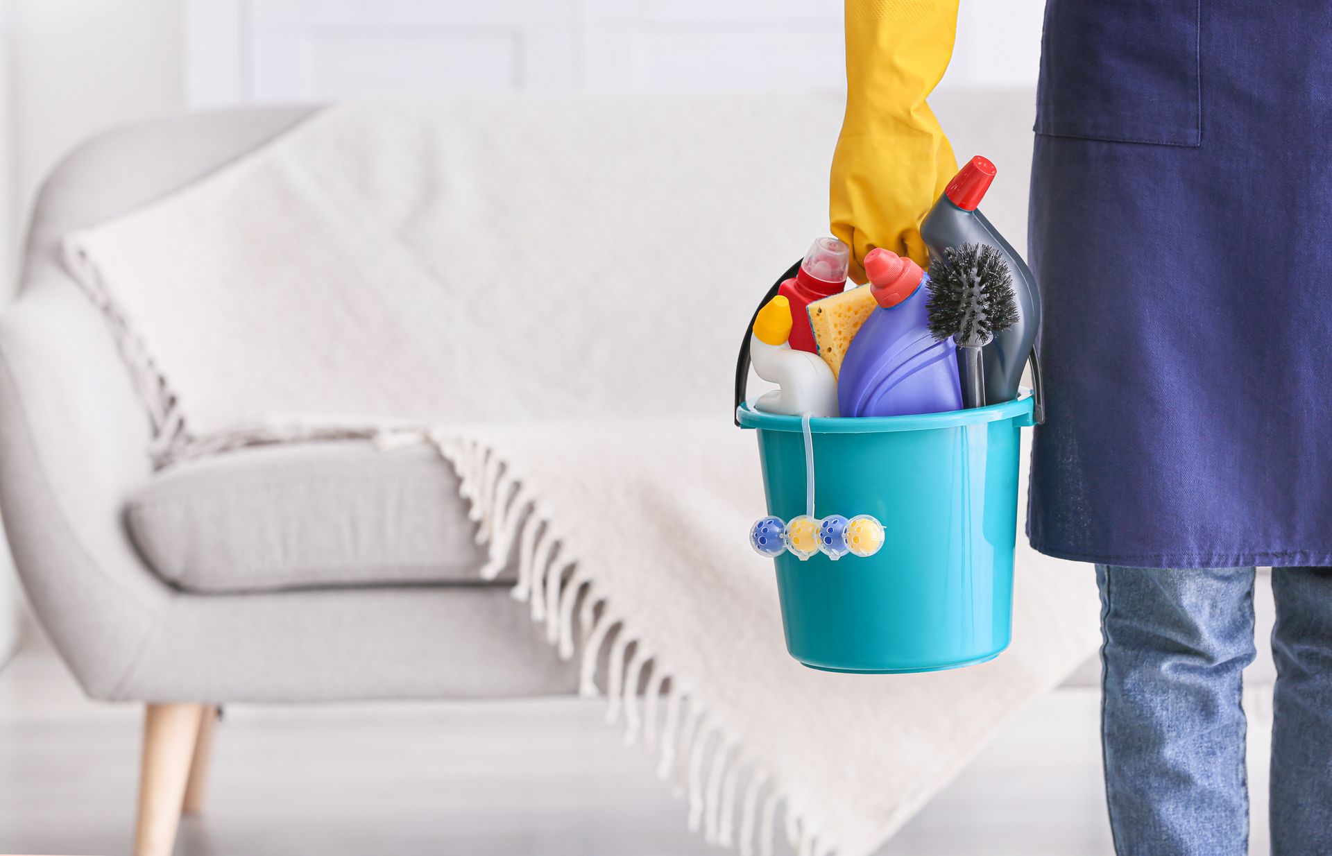 Person holding a blue cleaning bucket beside a couch in a bright living room
