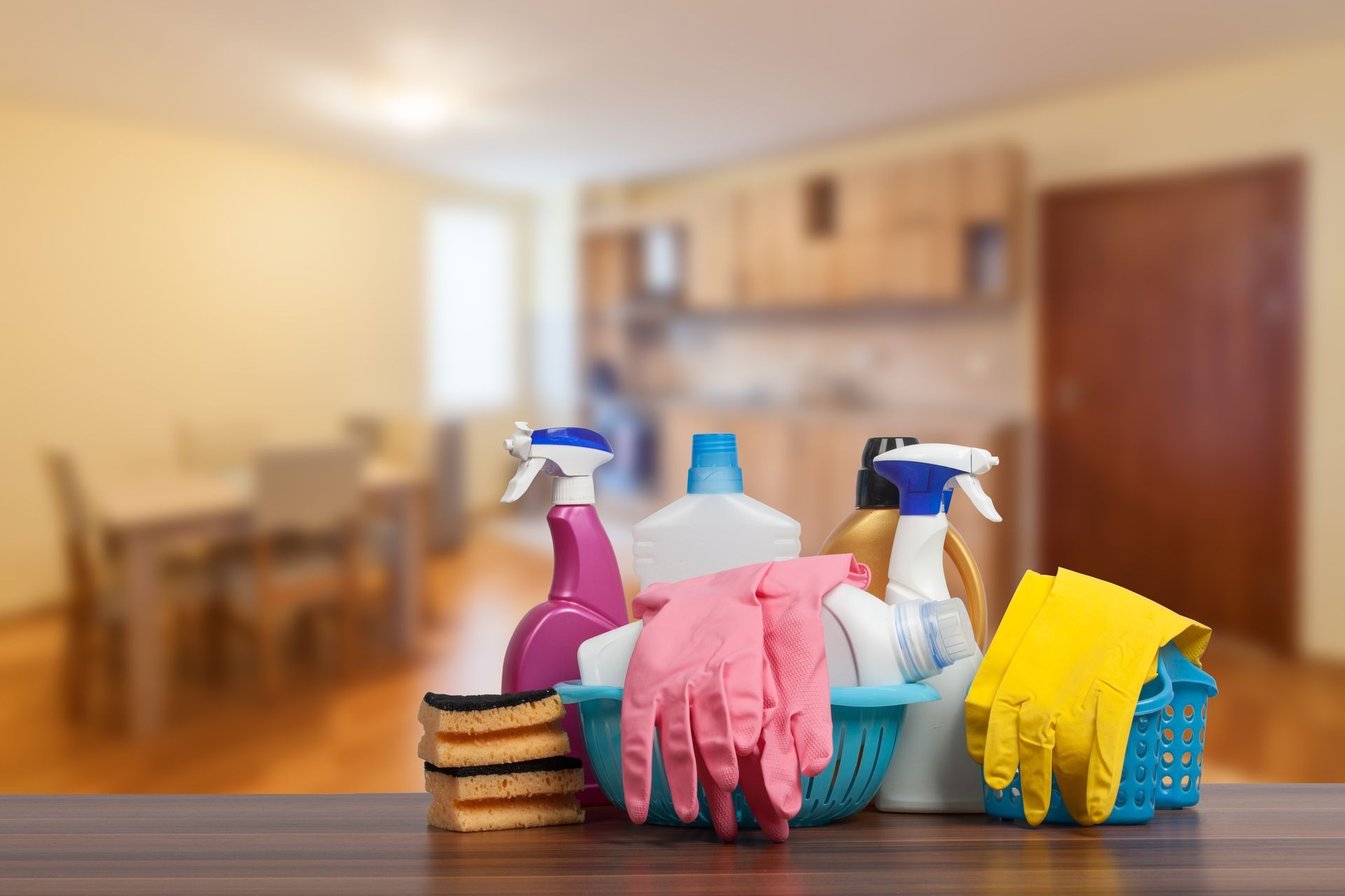 Cleaning supplies on a wooden table in a blurred kitchen, including spray bottles, gloves, and cloths.