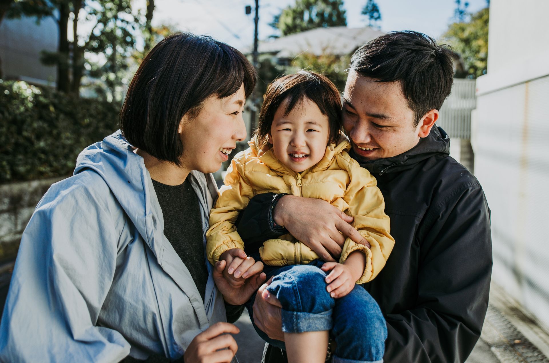 Smiling family of three outdoors, with a child in a yellow jacket being held between two adults