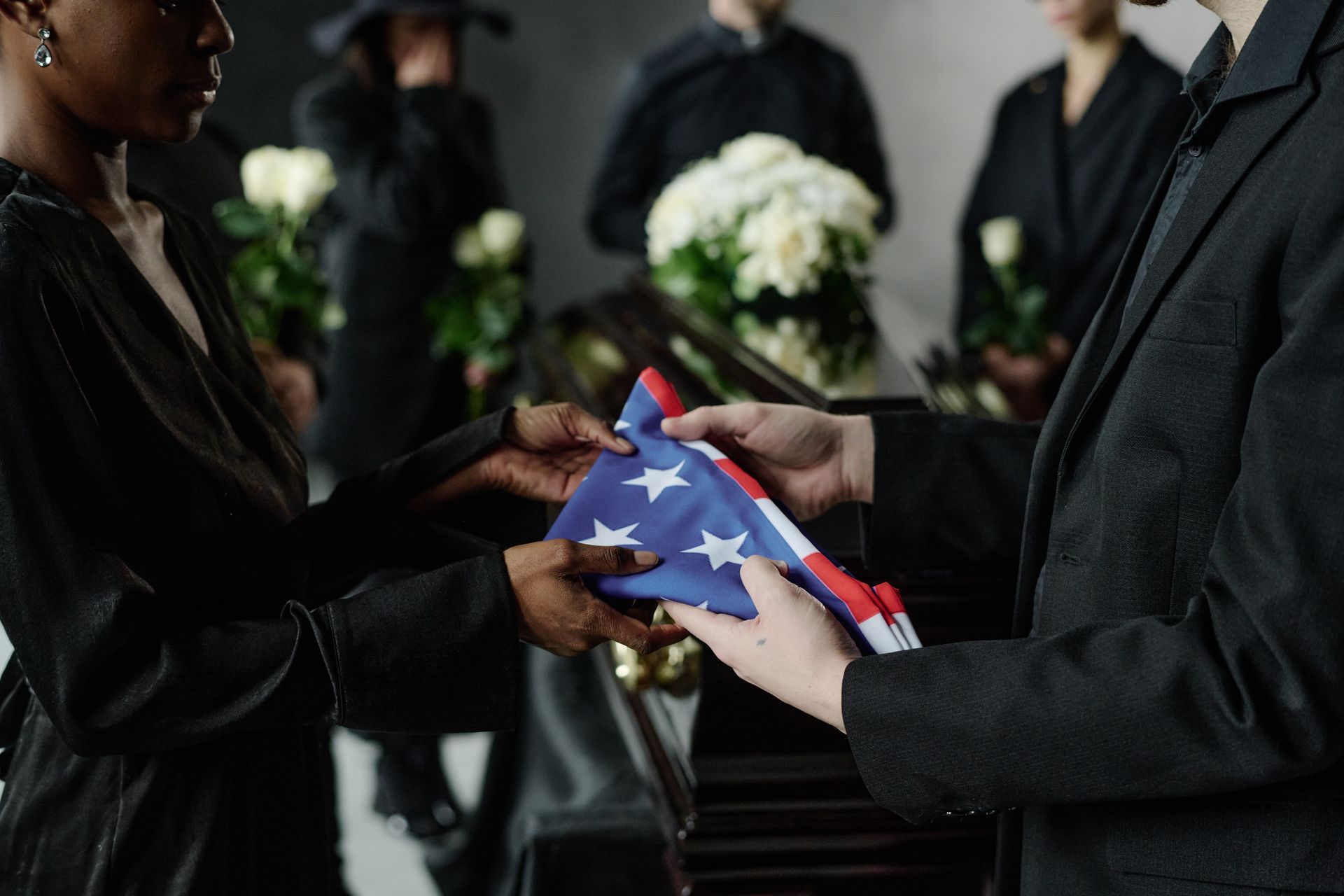 Person folding a folded U.S. flag at a funeral ceremony with mourners in black suits nearby