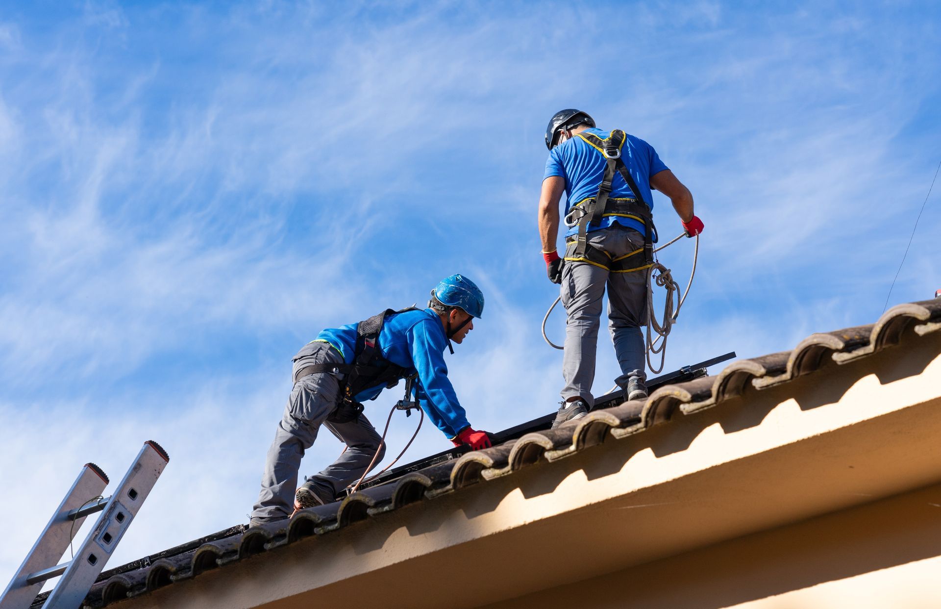 Two roofers in blue work on a tiled roof under a bright sky