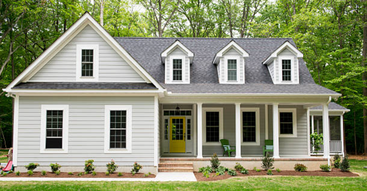 A white house with a yellow door and a porch surrounded by trees.