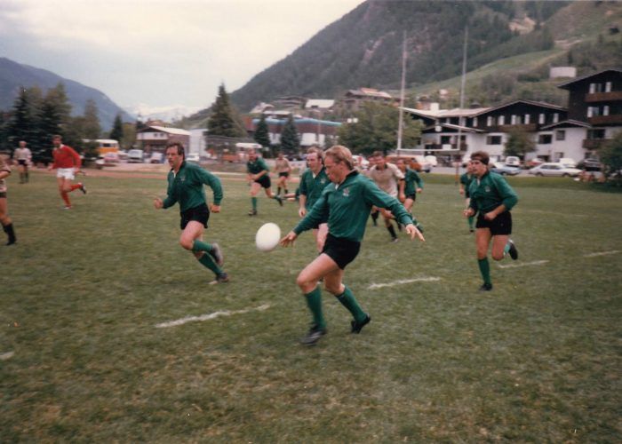 Players in green jerseys playing rugby on a field with mountains in the background, one player kicking the ball.