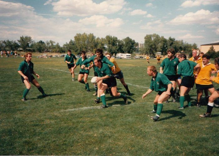 Rugby players in green and gold uniforms compete on a sunny grass field; one player in green carries the ball forward.