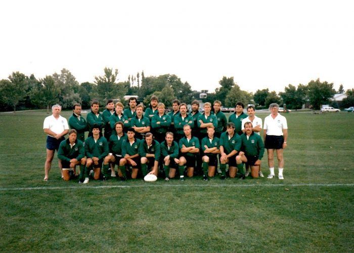 A rugby team in green jerseys and black shorts poses for a group photo on a grassy field with two coaches on the ends.