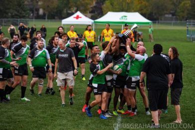 A rugby team in green and black uniforms celebrates on a grass field, lifting a trophy in the air.