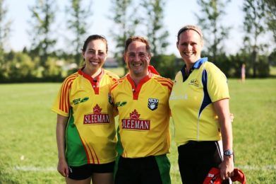 Three people wearing matching team shirts stand together smiling on a grassy field on a sunny day.