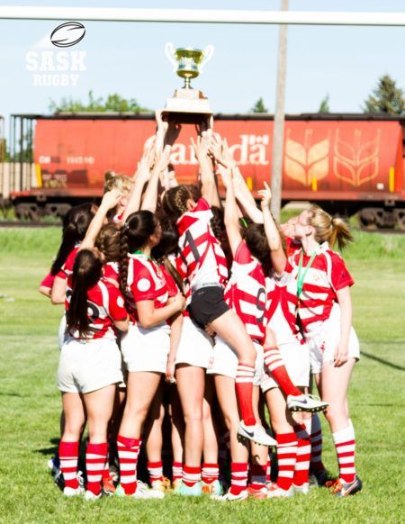 A rugby team in red and white uniforms lifts a trophy together on a field with a train in the background.