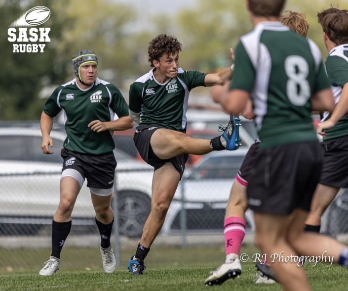 Rugby players in green uniforms on a field; one player kicks the ball while another runs nearby.