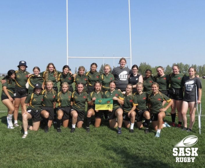 A team of rugby players in green and gold uniforms poses for a photo on a grassy field in front of rugby goalposts.