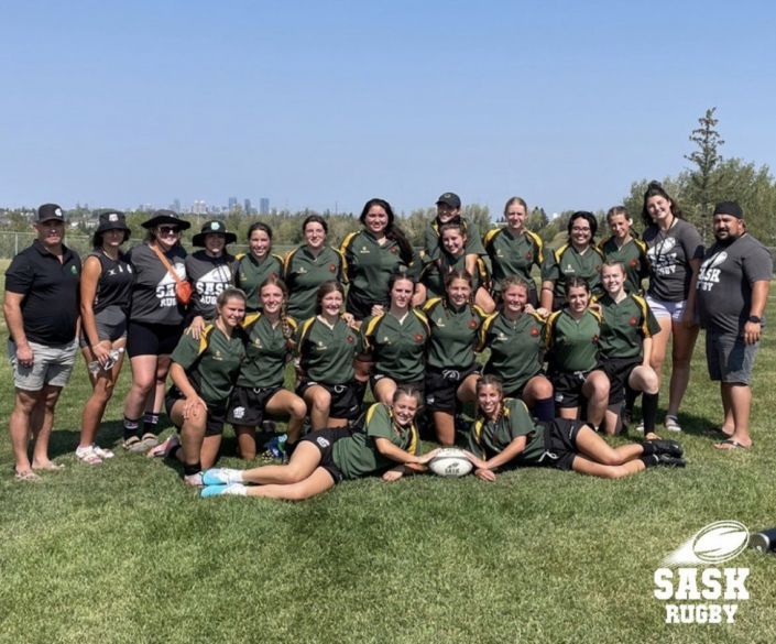 A team in green and black rugby jerseys poses on a field with two coaches, one player kneeling with a rugby ball.