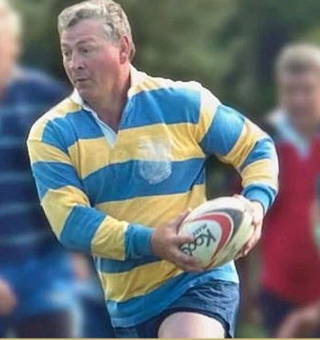 A person wearing a blue and yellow striped rugby shirt holds a rugby ball while running on a field.