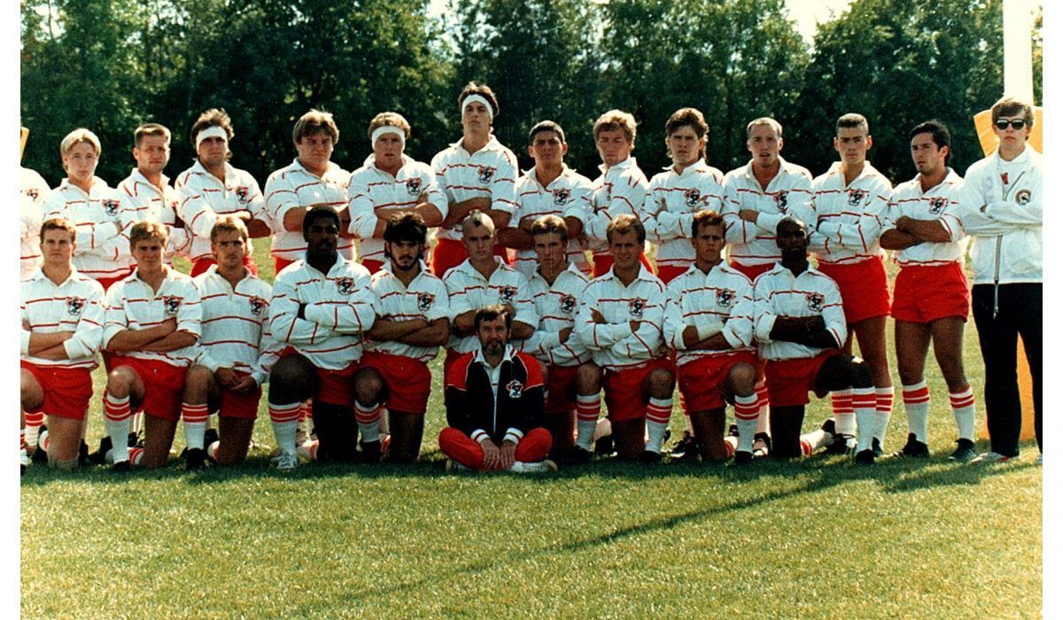 A rugby team in white jerseys and red shorts poses for a group photo on a grass field, with one player kneeling in front.