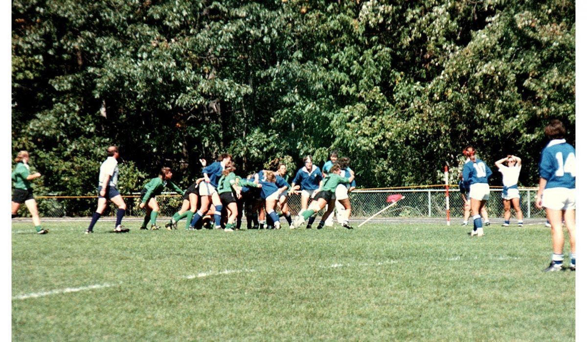 Rugby players in blue and green uniforms compete in a scrum on a grass field during a sunny match.