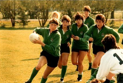 A group of athletes in green rugby jerseys run across a field, with one player in the foreground holding a ball.