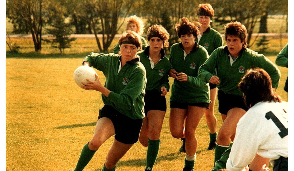 Players in green jerseys running on a field with a ball, while a teammate in a white jersey observes.