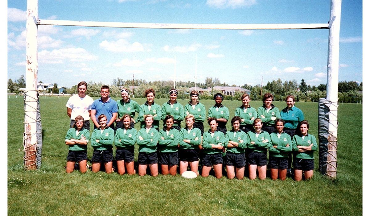 A rugby team poses in green jerseys and black shorts in front of goalposts on a grassy field with coaches standing behind.