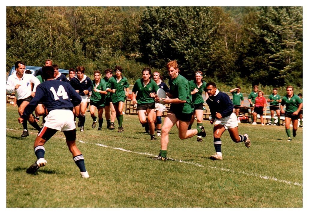 Rugby players in green and blue jerseys run across a grass field during a match, with one player carrying the ball.