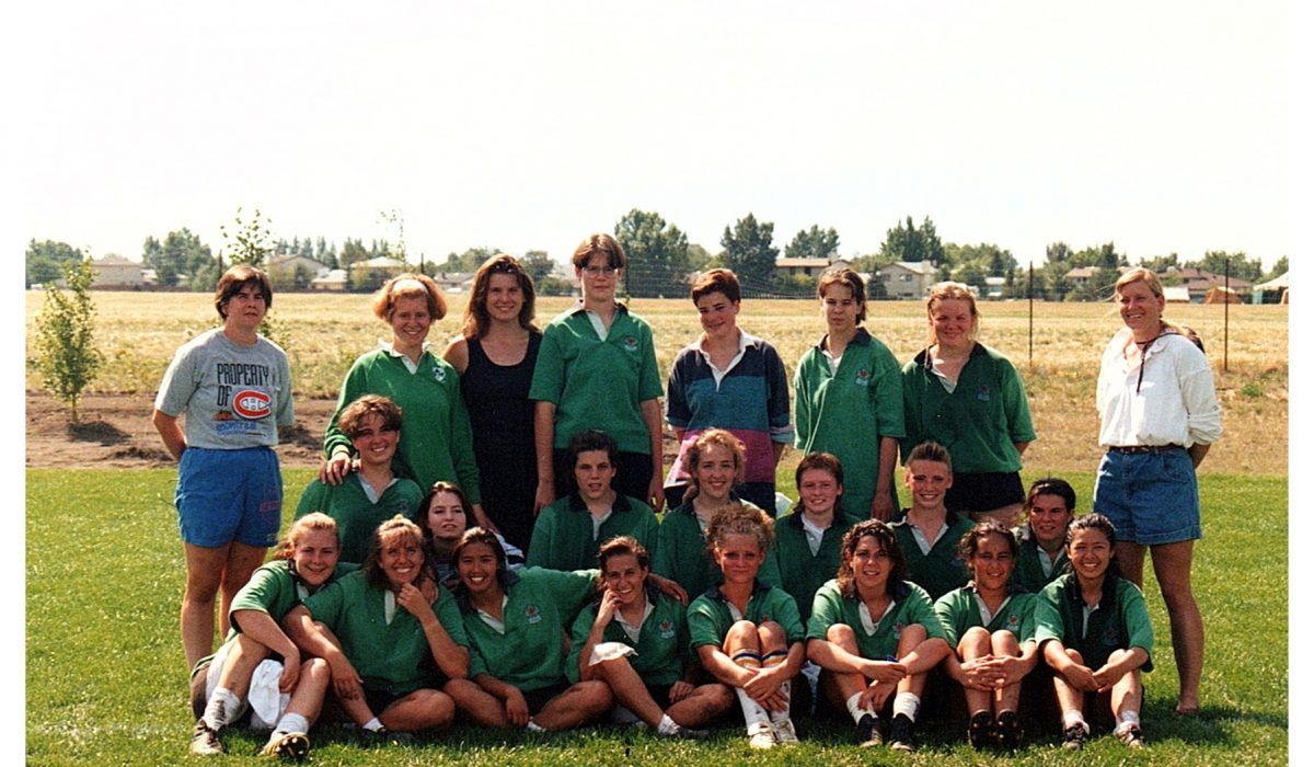 A group of people wearing green shirts and shorts pose for a team photo on a grassy field under a bright, sunny sky.