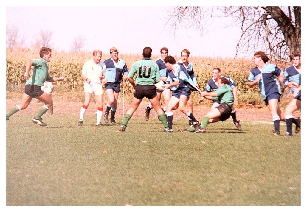 A group of athletes in green and blue rugby uniforms playing a match on a grass field near a field of corn.