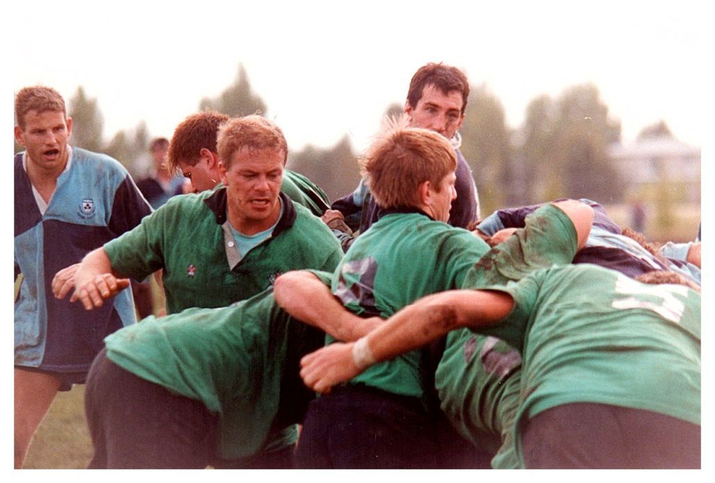 A rugby scrum in progress, with players in green jerseys engaged against players in blue and black jerseys outdoors.