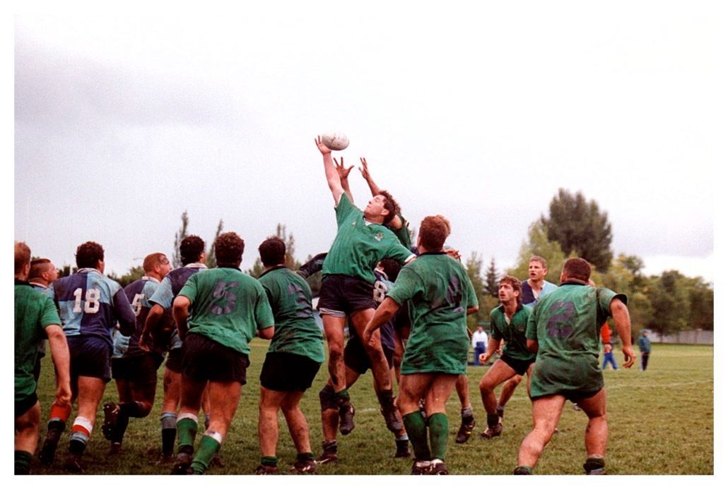 Rugby players in green and blue jerseys compete for a high ball during a line-out on an outdoor grass field.