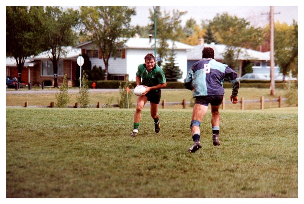 Two rugby players running on a grass field in front of suburban houses; one player carries the ball while the other follows.