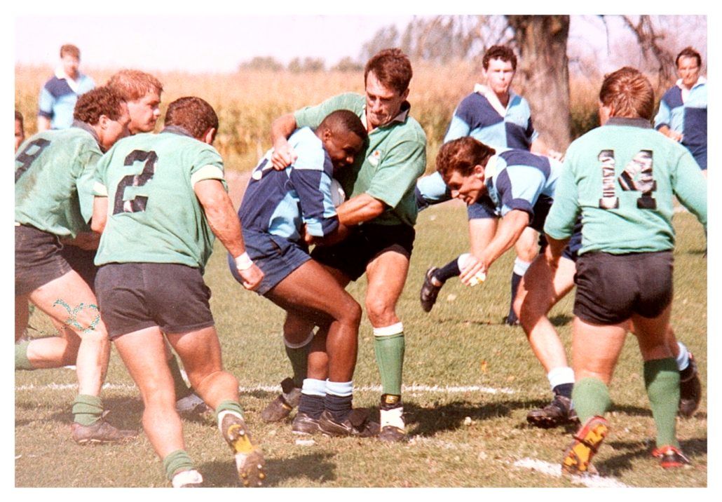 Rugby players in green and blue uniforms compete on a field, with one player in blue being tackled by two in green.