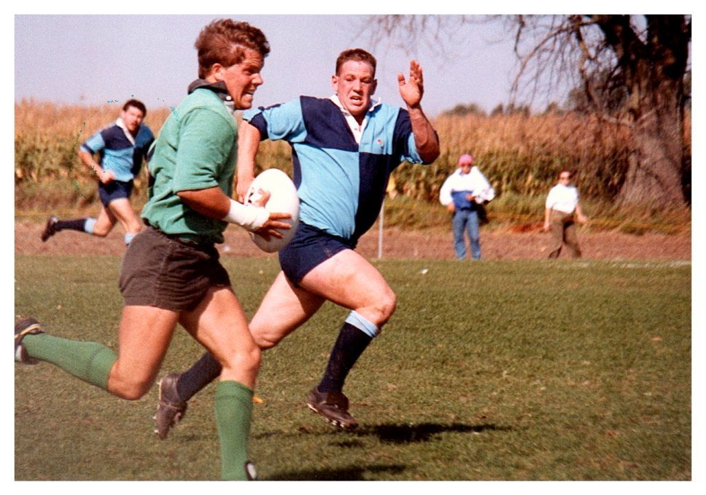 Two rugby players in team uniforms sprint across a field during a game, with others visible in the background.