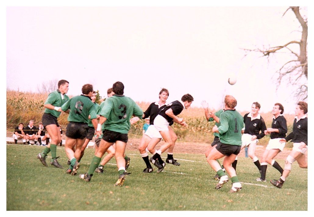 Players in green and black jerseys compete for a rugby ball during a match on a grass field with tall grass in the back.