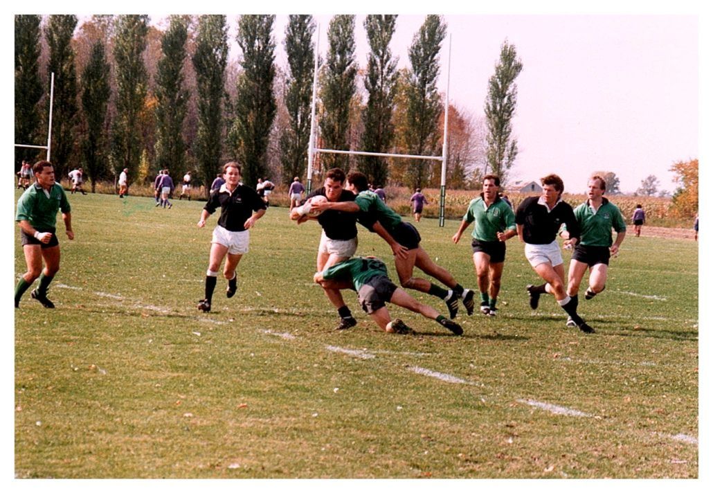A rugby match on a grass field, featuring players in green and black uniforms engaged in a tackle near the goalposts.
