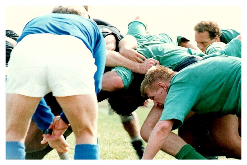 A group of rugby players in blue and green uniforms engaged in a scrum on a grass field.