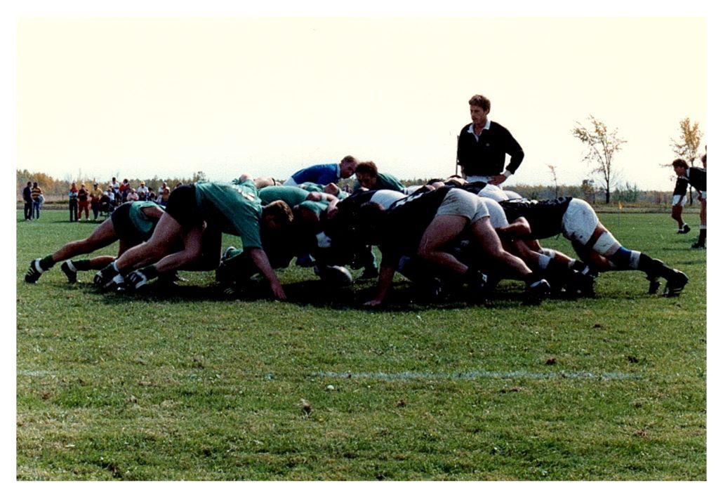 Rugby players in green and black uniforms engage in a scrum on a grass field while a coach observes nearby.