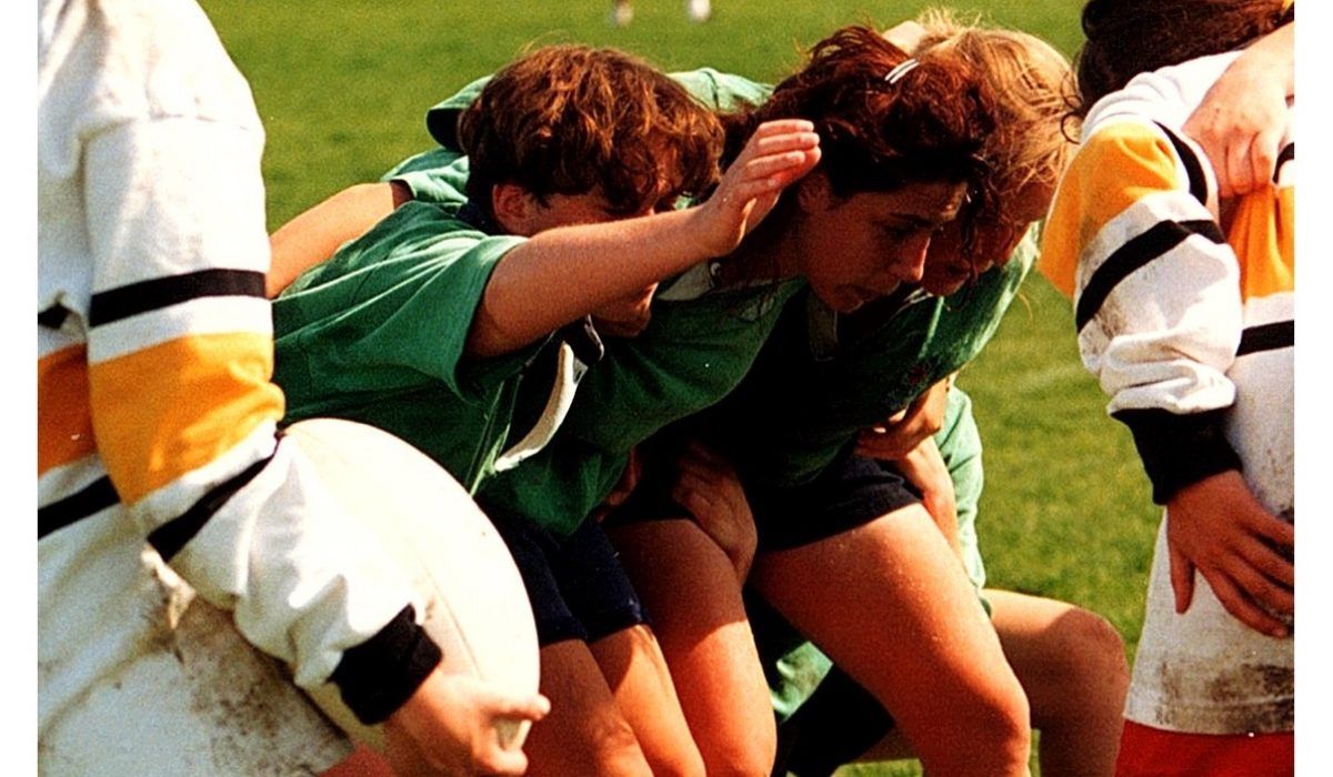 Players in green jerseys in a rugby scrum on a grass field, with players in yellow and white jerseys on either side.