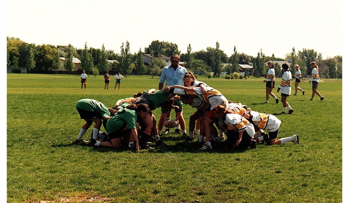 A rugby team in green and white jerseys forms a scrum on a grassy field, with a coach observing nearby.
