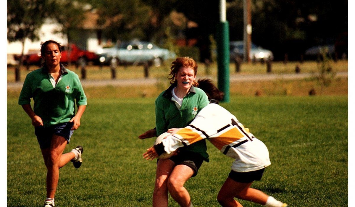 Three players in a rugby match on a grassy field; one in white tackles a player in green while another in green runs past.