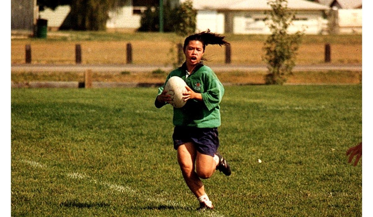 A person in a green jersey and dark shorts runs across a grassy field while holding a rugby ball.
