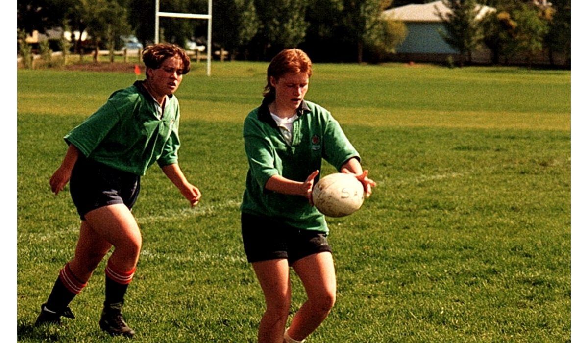 Two people in green rugby uniforms run across a grass field, one preparing to pass a rugby ball.