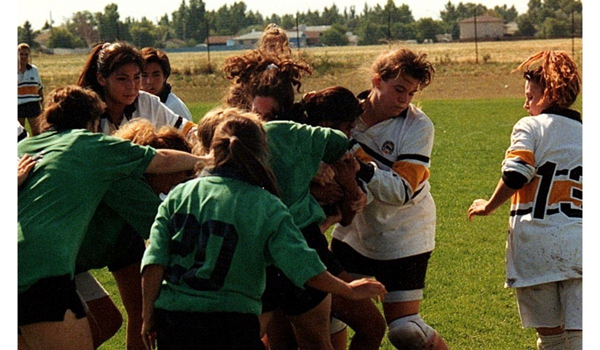 Players in green and white jerseys compete in a rugby scrum on a grass field during a sunny match.