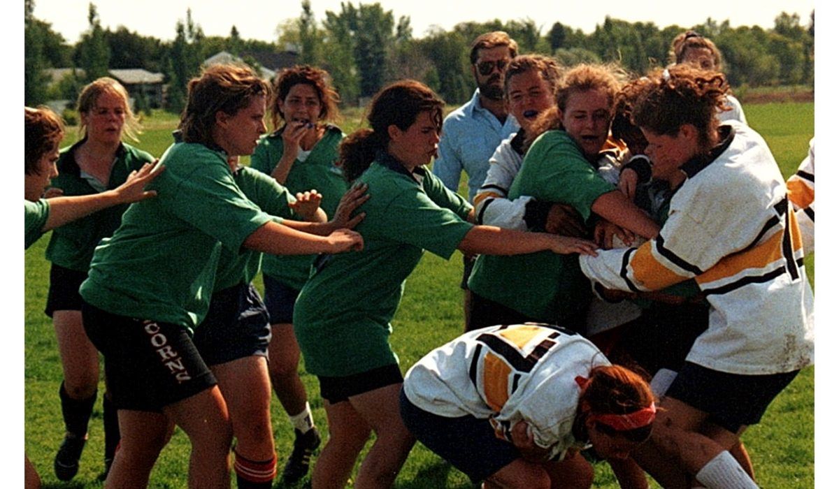 A rugby scrum on a grass field, with players in green and white/yellow uniforms engaging in a competitive match.