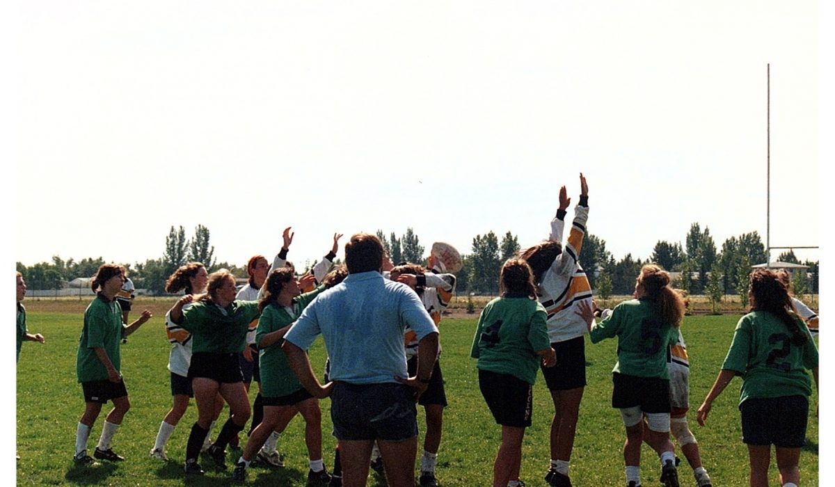A team in green and white uniforms plays rugby on a grass field, with players reaching up to catch a ball.