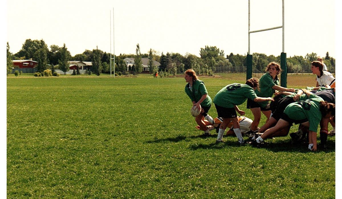 A rugby team in green uniforms engages in a scrum on a grassy field near a goalpost.