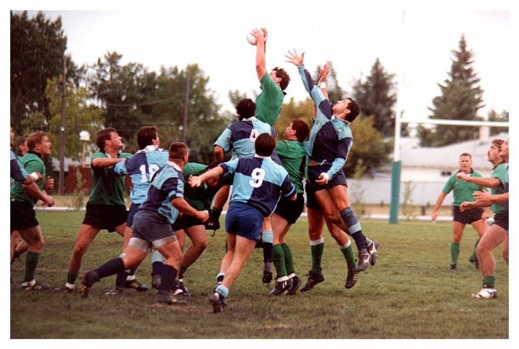 Rugby players in blue and green uniforms compete to catch the ball during a lineout on a grass field.