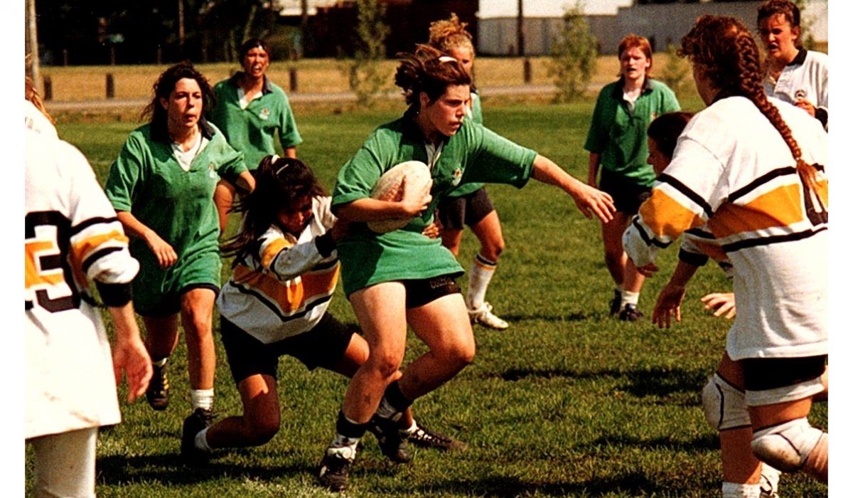 Players in green and white uniforms play a rugby match on a grassy field, with one player carrying the ball.