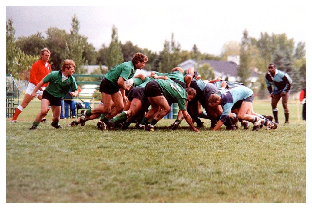 Rugby players in green and blue uniforms engage in a scrum on a grass field during a match.
