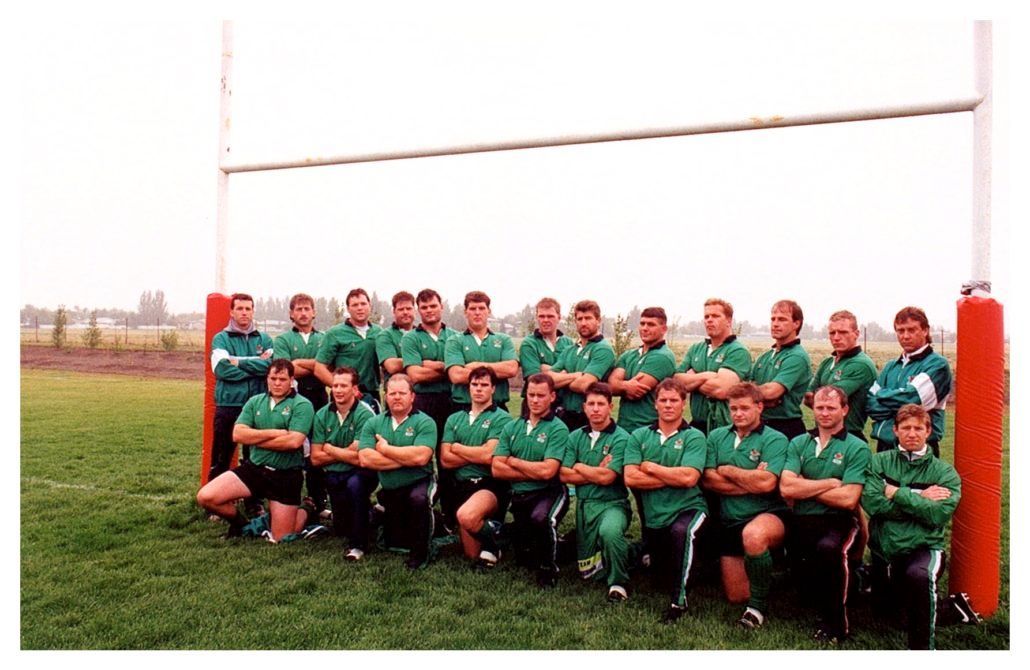 A group of rugby players in green uniforms posing for a team photo on a grassy field in front of rugby goalposts.